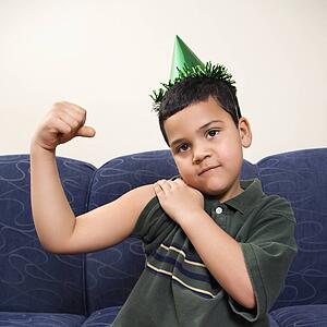 Boy wearing party hat playfully flexing arm muscle while looking at viewer