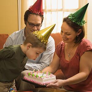Caucasian boy in party hat blowing out candles on birthday cake with family watching.