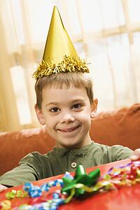 Caucasian boy wearing party hat holding gift and smiling at viewer.