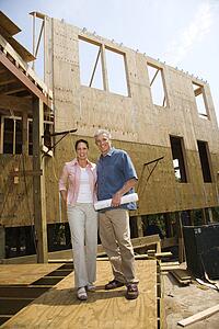 Caucasian mid-adult male holding blue prints with arm around mid-adult female in building construction site.