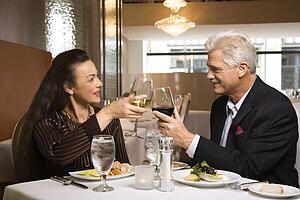 Caucasian mature adult male and prime adult female sitting at restaurant table toasting wine glasses.