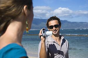 Man on beach pointing video camera at woman