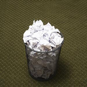Wire mesh trash can filled with crumpled paper on green carpet.