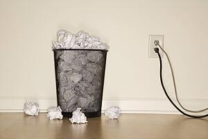 Full wire mesh trash can with crumpled paper next to electrical outlet and plugs.