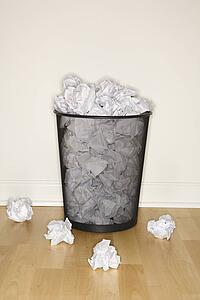 Wire mesh trash can filled and surrounded by crumpled paper.