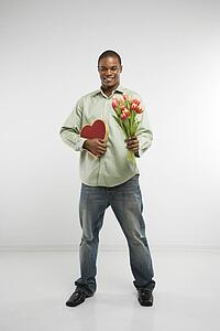 Studio shot of African-American man holding valentine heart and bouquet of tulips smiling and looking at viewer.