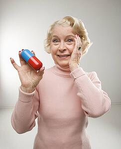 Caucasian senior woman holding oversized pill at viewer.