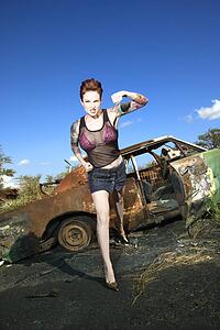 Sexy tattooed woman standing with defiant look punching in front of old rusted car in junkyard