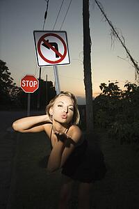 Caucasian mid-adult blonde woman wearing little black dress blowing kiss at viewer beside no left turn sign and stop sign.