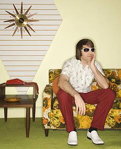 Caucasian mid-adult man wearing sunglasses sitting on colorful retro sofa looking bored.