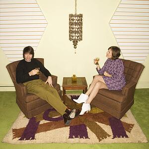 Caucasian mid-adult man and woman wearing vintage clothing seated in brown retro chairs smiling and drinking.