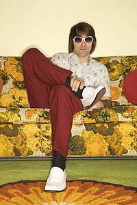 Caucasian mid-adult man sitting on colorful retro sofa wearing sunglasses looking bored.
