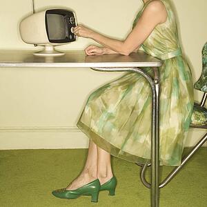 Side view of Caucasian mid-adult woman wearing green vintage dress sitting at 50's retro dinette set turning old televsion knob.