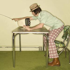 Side view of Caucasian mid-adult man wearing hat and plaid pants leaning over 50's retro dinette set adjusting old television set.