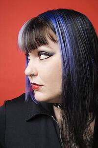 Portrait of woman with blue hair, tattoo, and spike collar looking to side against orange background