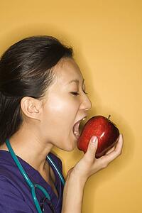 Asian Chinese mid-adult female doctor taking bite of red apple against yellow background.