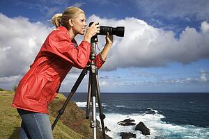 Woman looking through camera on tripod on cliff overlooking ocean in Maui, Hawaii