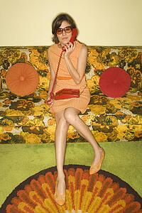 Pretty Caucasian mid-adult woman wearing sunglasses sitting on colorful retro sofa talking on red telephone.