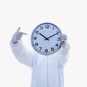 Man in biohazard suit holding clock in front of face pointing to clock with finger standing against white background.