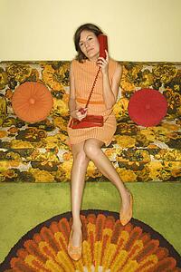 Pretty Caucasian mid-adult woman sitting on colorful retro sofa talking on red telephone.