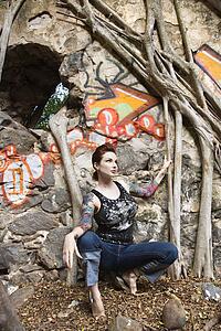Sexy Caucasian tattooed woman crouching next to wall covered in graffiti and Banyan tree branches.