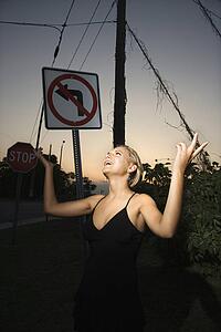 Caucasian mid-adult blonde woman wearing little black dress posing with hands up beside no left turn sign.