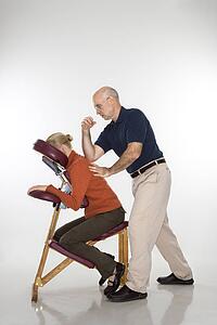 therapist massaging a woman's back sitting in massage chair with his elbow. 