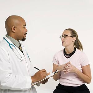 Mid-adult African-American doctor with clipboard and Caucasion mid-adult female patient in doctor's office looking at viewer and smiling.