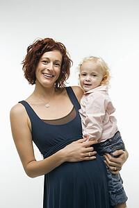 Portrait of Caucasion mid-adult attractive pregnant woman standing, holding female toddler on hip and other hand on belly, looking at viewer and smiling.