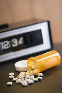 Open medicine bottle with pills spilling out onto table with clock in background.