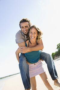 Mid-adult Caucasian woman giving man piggyback ride on beach. 