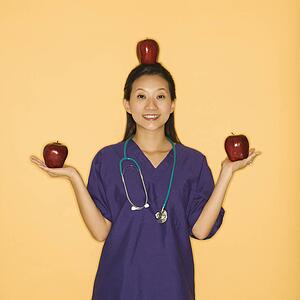 Asian Chinese mid-adult female doctor holding two red apples in hands and balancing one on head against yellow background smiling and looking at viewer.
