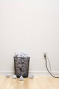Full wire mesh trash can with crumpled paper next to electrical outlet and plugs