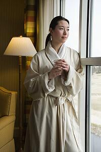 Woman in bathrobe looking out window holding cup.