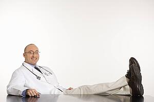 Caucasian mid adult male physician sitting with feet on desk smiling.