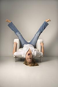 Mid-adult woman in blue jeans lying upside down on chair with legs in air laughing