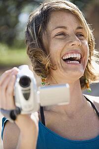 Mid-adult Caucasian woman outdoors holding video camera laughing.