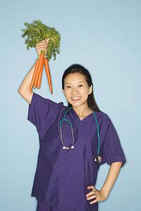 Asian Chinese mid-adult female doctor with hand on hip holding up bunch of carrots against blue background smiling and looking at viewer.