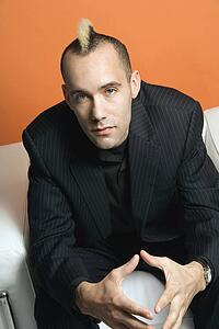 Caucasian man in suit with mohawk sitting in chair against orange background.