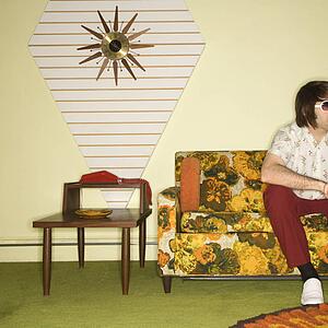 Caucasian mid-adult man sitting on colorful retro sofa wearing sunglasses in room with vintage decor.