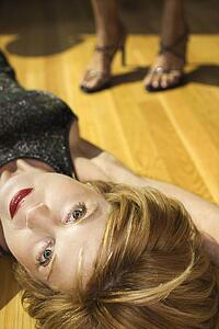 Adult woman lying on wood floor looking at viewer with feet of another woman in background wearing heels.