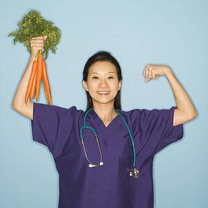 Asian Chinese mid-adult female doctor flexing muscle holding up bunch of carrots against blue background smiling and looking at viewer.