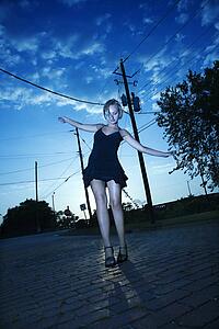 Caucasian mid-adult blonde woman wearing little black dress posing in street at dusk.