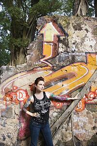 Sexy tattooed Caucasian woman standing next to wall covered in graffiti and a tree branch.
