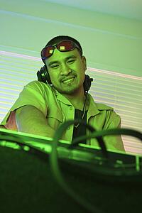 Young adult DJ behind mixing equipment looking at viewer smiling