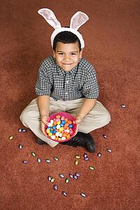 Hispanic boy wearing bunny ears sitting on floor holding a bowl of chocolate candy eggs with more candy scattered around him looking up at viewer smiling.