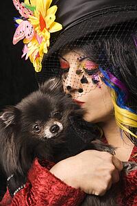 Caucasian woman in unique makeup and clothing holding and kissing black Pomeranian dog.