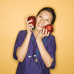 Asian Chinese mid-adult female doctor holding two red apples up to face against yellow background smiling and looking at viewer.