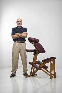 Male massage therapist standing with arms crossed beside massage chair