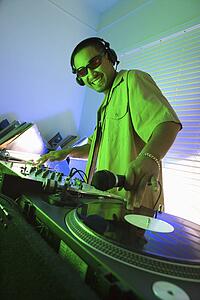 Young adult DJ wearing sunglasses behind mixing equipment with hand on record turntable looking at viewer smiling